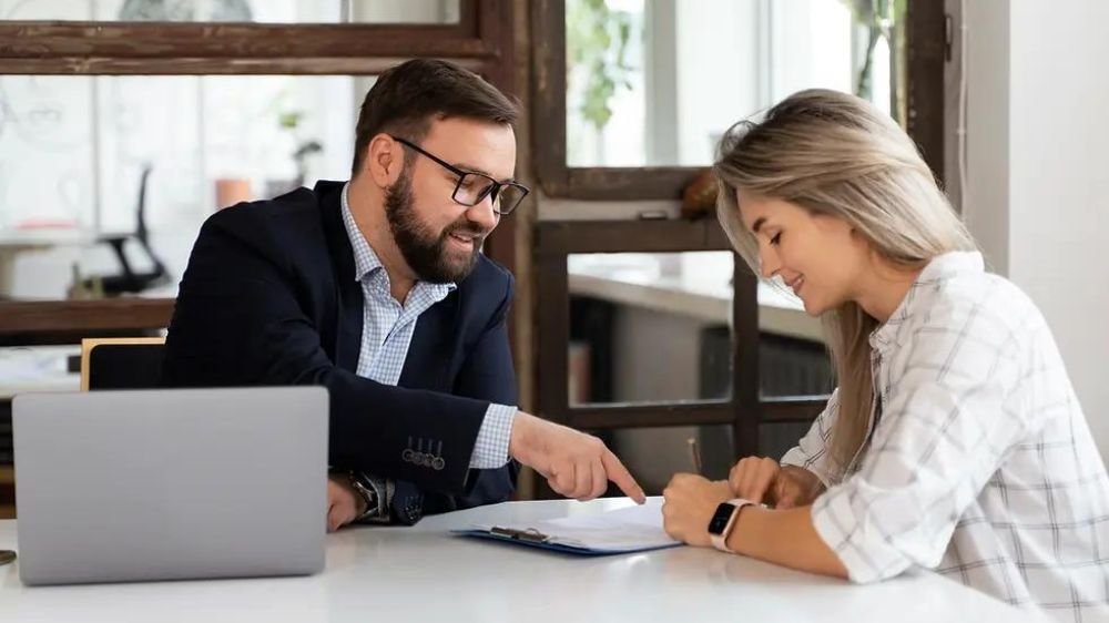 business-insurance-application-consultation Business professional discussing paperwork with a woman who is completing documents at a desk