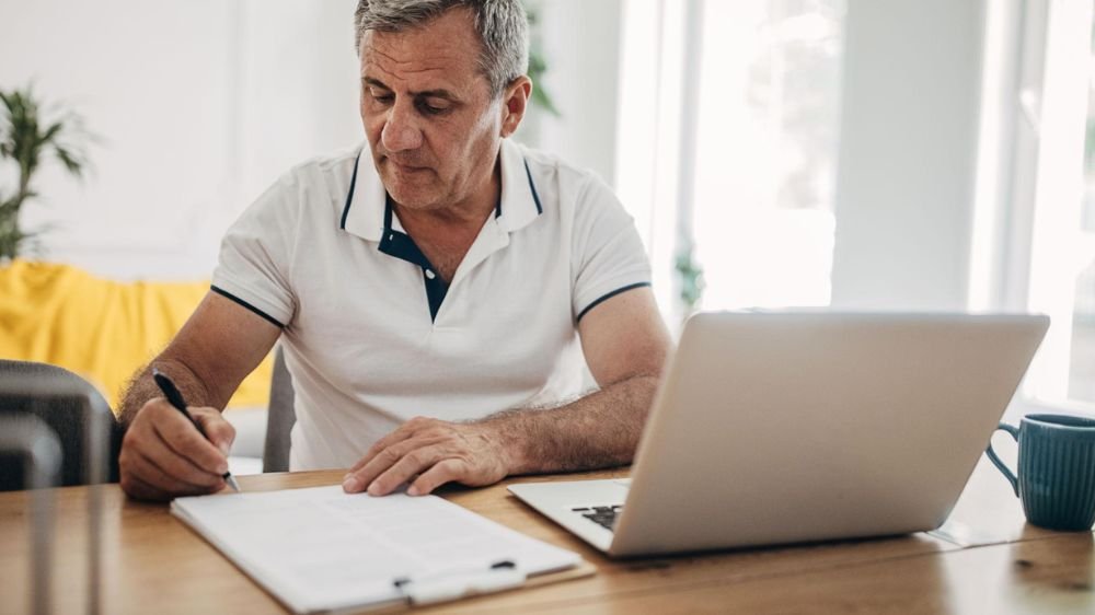 business-owner-reviewing-license-documents Business owner writing on paperwork while working on a laptop at his desk.