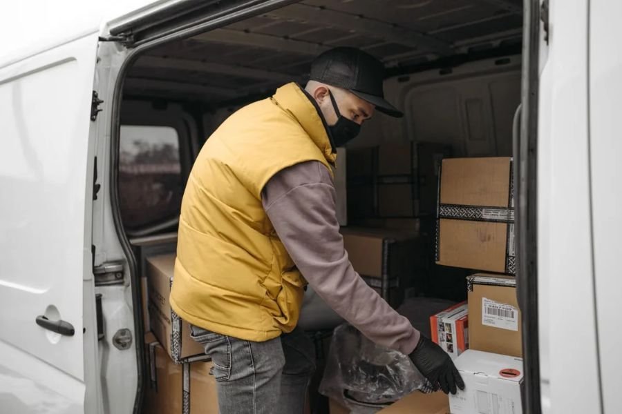 delivery-worker-handling-cargo-packages Delivery worker wearing a yellow vest and face mask handling cardboard packages inside a white cargo van
